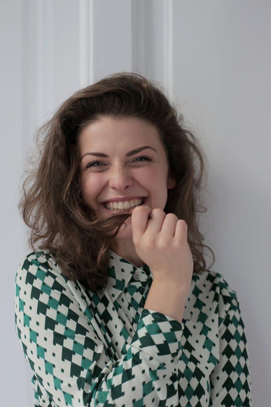 happy young woman touching hair while standing near white wall